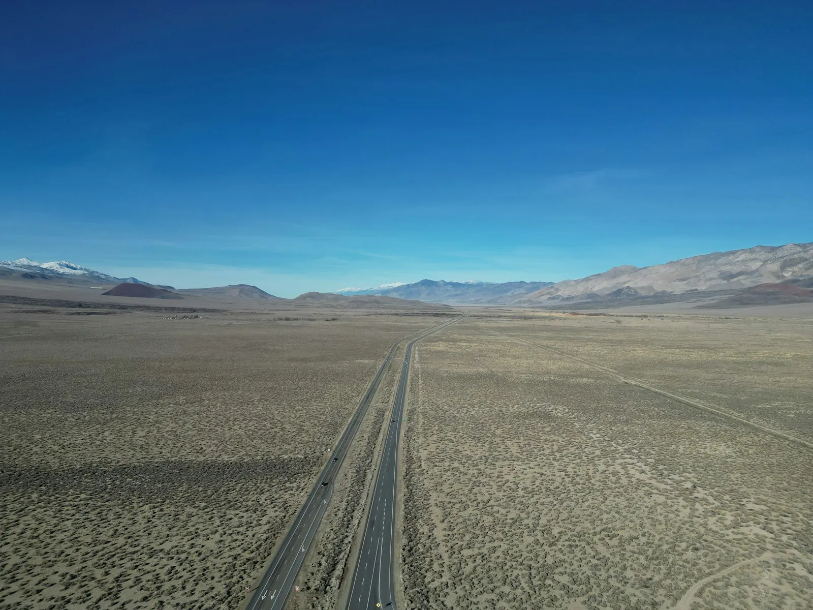 A lone highway crossing an empty California high-desert basin, snow-capped Sierra Nevada in the distance.